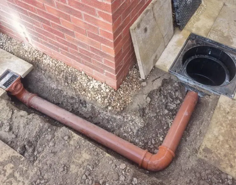 New orange drainage pipes being laid underground near a brick wall and patio surface, showing the connection points and an open inspection chamber.