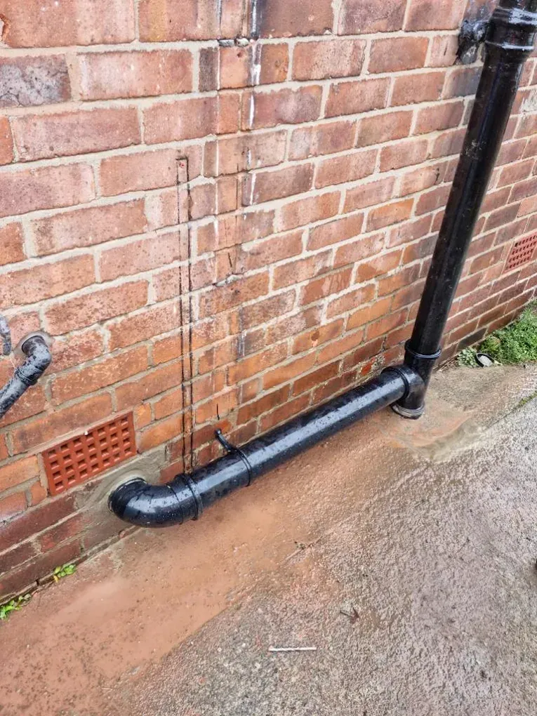 Blocked drain pipe with water pooling outside a brick building wall.